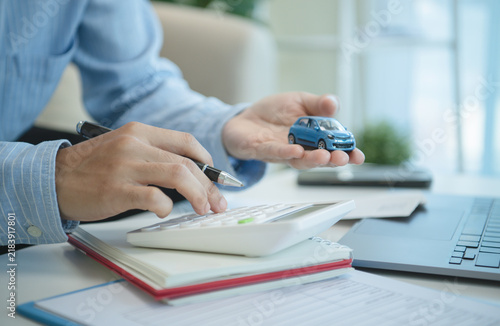 Business professional holding model car and using calculator beside laptop for auto loan planning vehicle financing insurance budgeting ownership costs and smart purchase decisions.