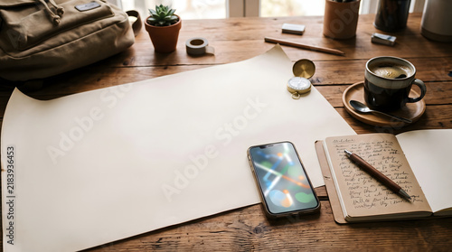 Blank paper sheet lying on rustic wooden desk surrounded by traveler gear including compass coffee phone and leather bound journal