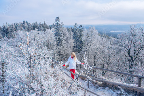 Woman in red pants and white jacket walking on a frosty wooden path in winter forest