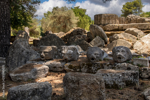 Fotografía "The image captures the sunlit archaeological ruins of Olympia, featuring ancient stone columns and weathered remnants scattered across a green landscape under a bright sky