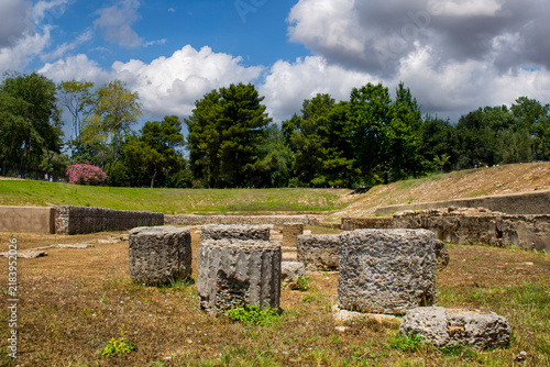 Foto "The image captures the sunlit archaeological ruins of Olympia, featuring ancient stone columns and weathered remnants scattered across a green landscape under a bright sky