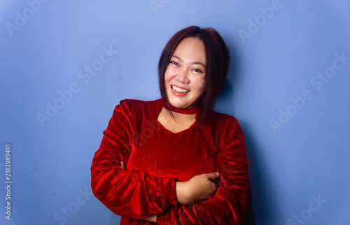 Cheerful asian woman in red dress leaning slightly with arms crossed on blue background