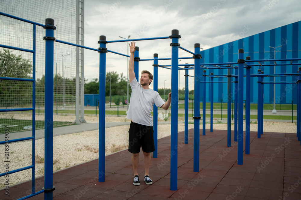 Obraz premium Man stands at outdoor fitness area with blue bars and gray sky, preparing for workout while looking around