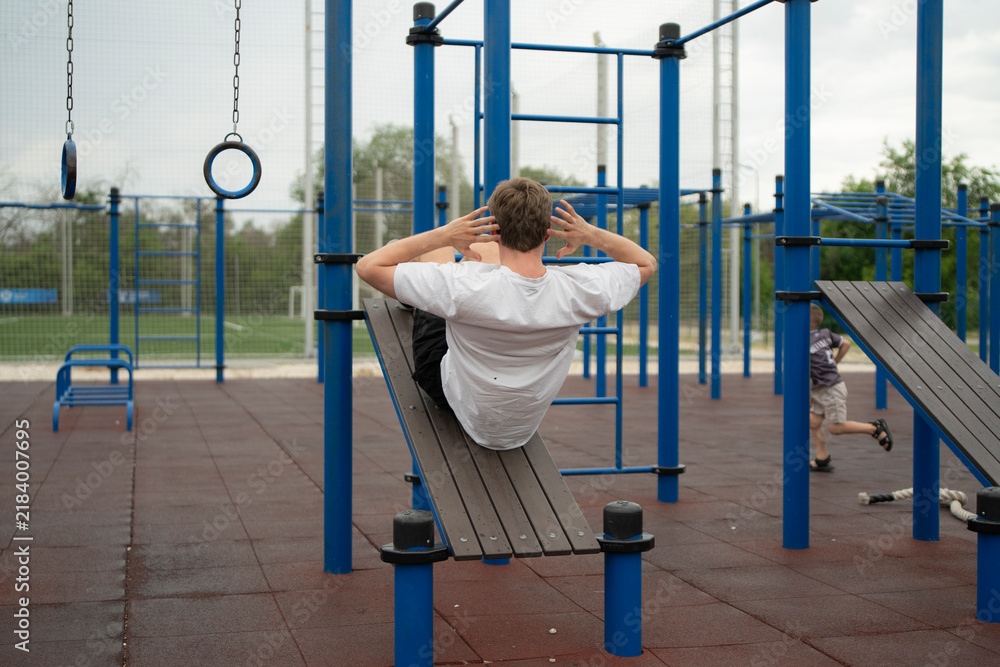 Fototapeta premium A man performs an ab workout on a bench in a fitness area while a child plays nearby in a park