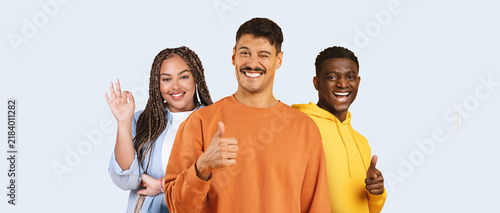 Fotografie Happy Multiracial Group Of Three Friends, Two Women And One Man, Smiling And Showing Thumbs Up With Positive Emotion, Standing Together In Studio Against Light Background