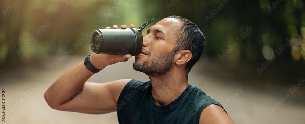 Fototapeta premium Happy Young Man Enjoying Hot Beverage From Black Thermos While Relaxing Outdoors, Focusing On His Wellbeing During Fitness Activities In A Natural Setting.