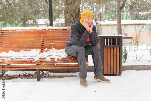 A 40-year-old man sits thoughtfully on a snow-covered wooden park bench in winter, wearing a jacket and yellow beanie.