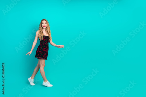 Фотография Young chic woman in a black red polka dot dress walks on teal background for lif