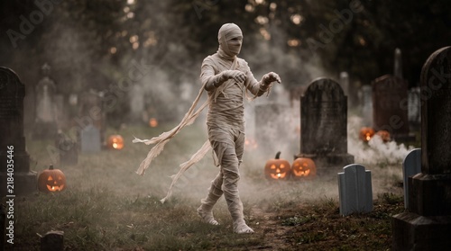 Mummy walking in foggy cemetery with glowing pumpkins at Halloween