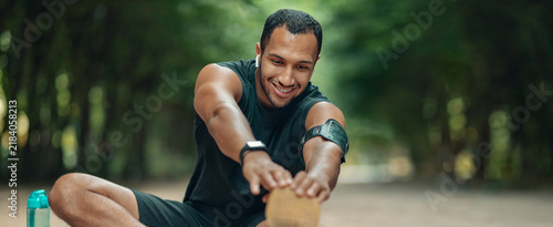 Konstfotografi Smiling Black Male Athlete Stretching On Pathway In Lush Green Outdoor Environment, Engaging In Warm-up Routine To Maintain Fitness, Showcasing Healthy Lifestyle And Wellbeing Concept