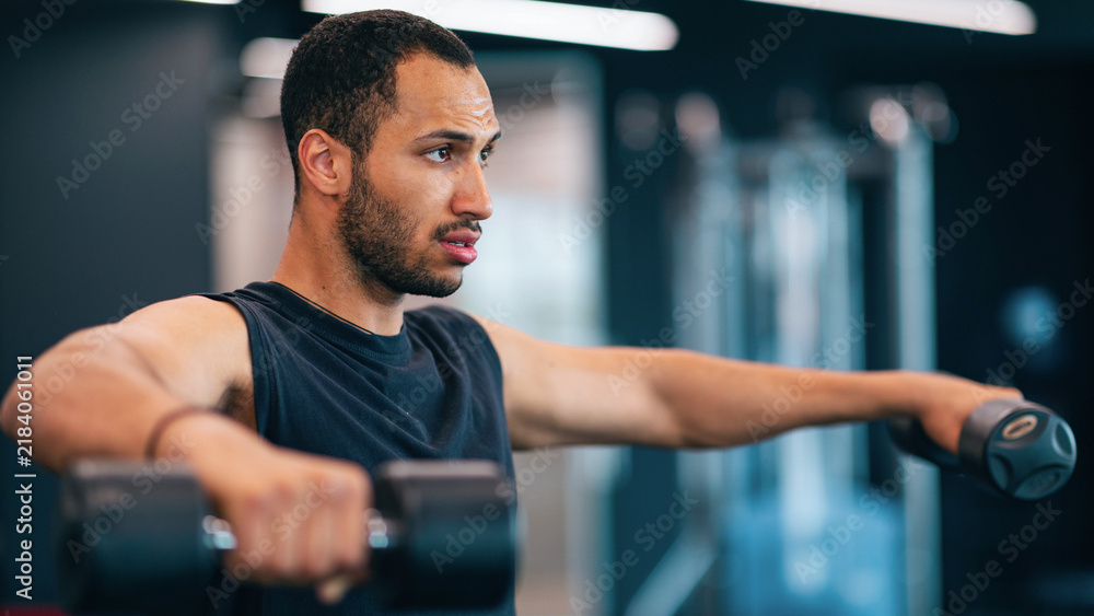 Fototapeta premium Focused Middle Eastern Man Performing Dumbbell Lateral Raises In Gym, Strength Training Session. He Is Engaged In Exercise Routine That Enhances Muscle Strength And Fitness.