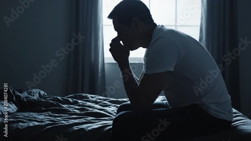 Man sitting on a dark bed in a hunched position with clasped hands, showing despair and emotional exhaustion.