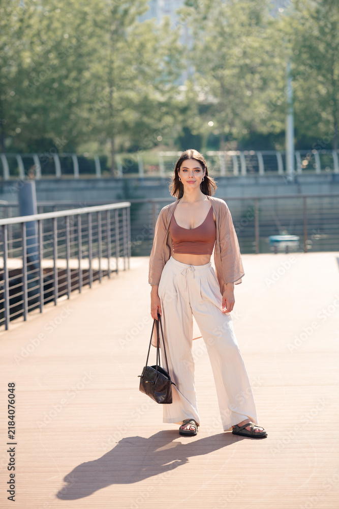Fototapeta premium Young woman in casual summer fashion walking on urban boardwalk with handbag — relaxed confident portrait in sunny city park