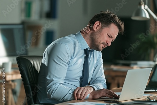 Stressed businessman working late at office desk with laptop while experiencing neck pain