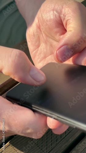 Man holding black smartphone with both hands to text friends online. Adult person typing message on dark mobile screen sitting at outdoor table. Digital connection and messaging via modern