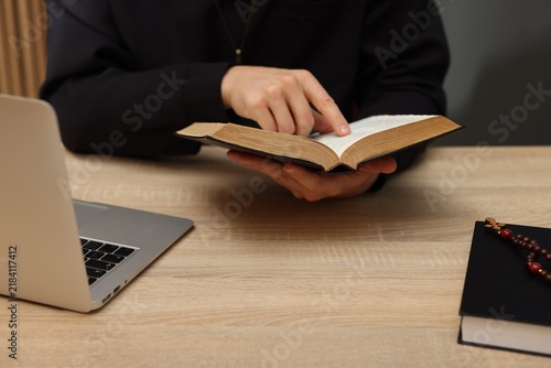 Принти на полотні Priest in cassock reading Holy Bible at wooden table indoors, closeup