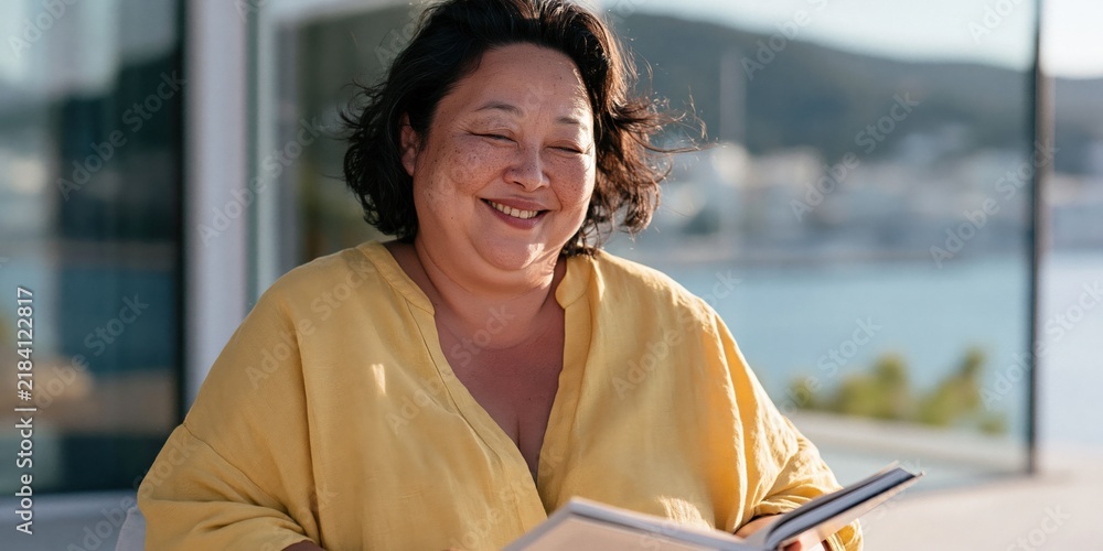 Obraz premium Joyful woman reading a book on a terrace by the sea for relaxation background and coastal lifestyle content