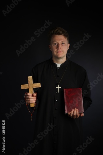 Obraz na plátne Priest in cassock with wooden cross and Holy Bible on black background