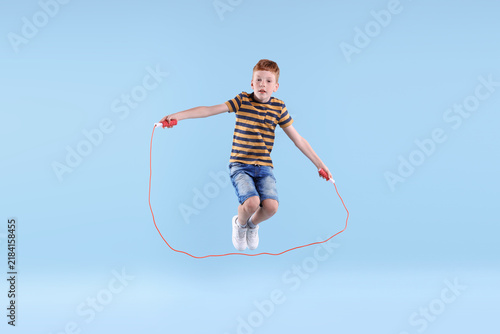 Cute little boy jumping with skipping rope on light blue background