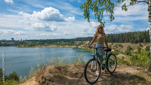 A girl stands next to her bicycle by a lake surrounded by trees. The sun shines brightly in the sky. The scene shows a clear day with few clouds and a green landscape.