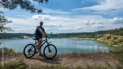 A man is riding a bicycle along a path next to a lake. The scene includes trees and hills in the background. The sun shines brightly in the sky. It is a clear day.