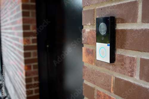 Smart doorbell camera mounted on red brick exterior wall beside residential front door in daytime, showing illuminated ring button, lens, and security monitoring