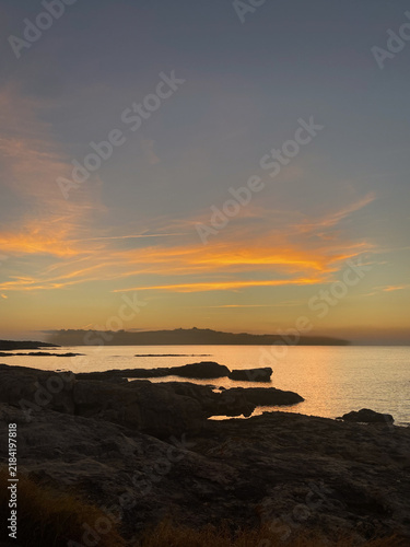 Landscape of rocky coast in Porto Vecchio, Corsica, France, during a warm golden hour sunset. The calm sea reflects orange sky, with silhouette Punta de la Chiappa, 