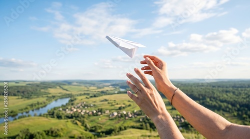 Hands launching paper airplane over sunlit countryside with river and blue sky