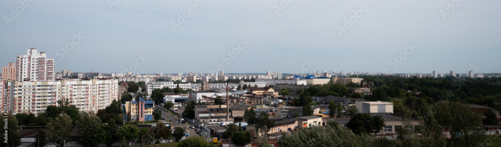 Obraz premium Wide urban skyline with apartment blocks, low-rise buildings, trees, and distant city horizon under soft cloudy sky, showing dense residential districts and calm summer atmosphere