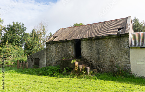 Old stone barn with a corrugated tin roof sitting under a partly cloudy sky. The historic, weathered outbuilding features an upper loft entrance and concrete steps on an Irish country farm.