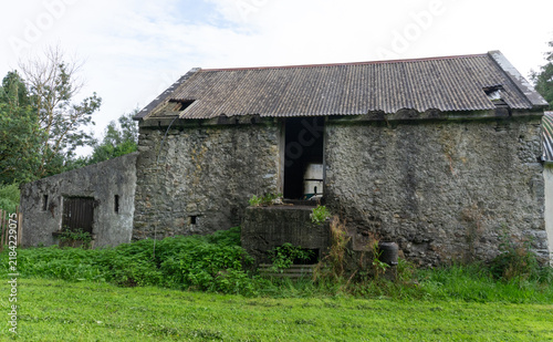 An old stone barn with a weathered corrugated iron roof stands on an abandoned Irish dairy farm. The rough stone walls feature a raised loft doorway, surrounded by green grass and overgrown weeds.