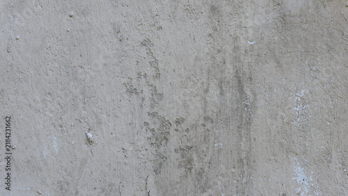 Texture of a weathered concrete wall. The flat, gray surface shows signs of age with subtle cracking, flaking plaster patches, dark dirt streaks, and a gritty industrial finish.