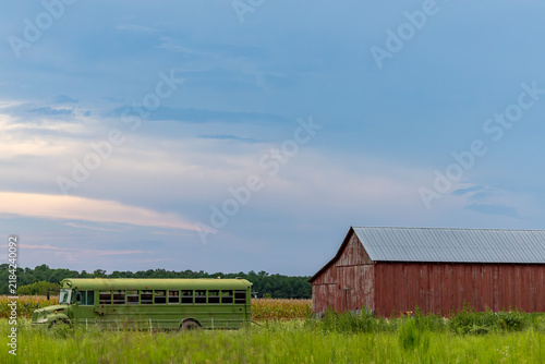 St. Mary's City, Maryland USA An abandoned green school bus on a cornfield with a cloudscape and a red barn.