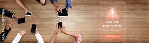 Фотография A group of individuals using various devices on a wooden table while an alert for a suspicious login attempt appears, highlighting digital security and teamwork in a modern workspace