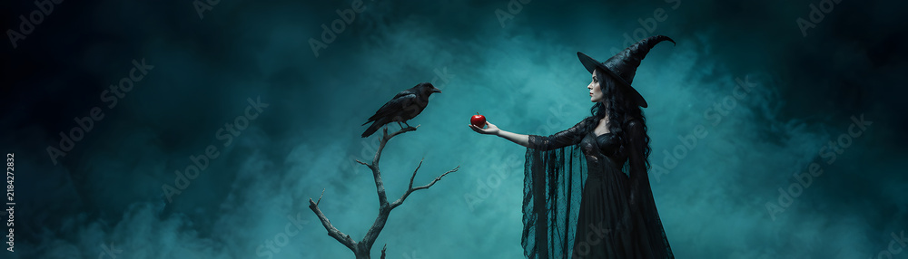 Obraz premium Mysterious witch stands in dark forest holding poison apple while raven watches closely, creating enchanting Halloween scene