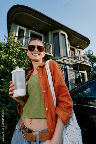 Smiling young woman in sunglasses and orange shirt holds a water bottle near a modern house. Summer lifestyle concept of refreshment  travel and healthy living outdoors.
