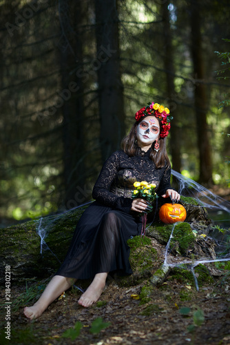 Young barefoot woman in the forest on a stump with a halloween pumpkin and yellow roses in hands.