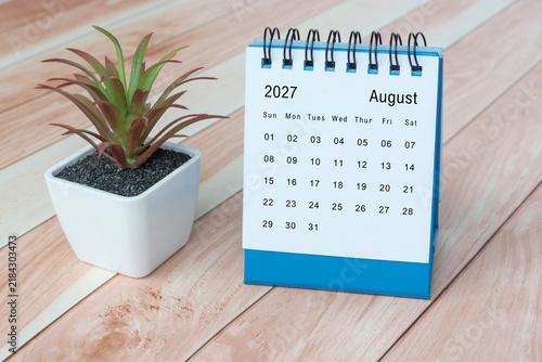 August 2027 desk calendar with potted plant on wooden desk.