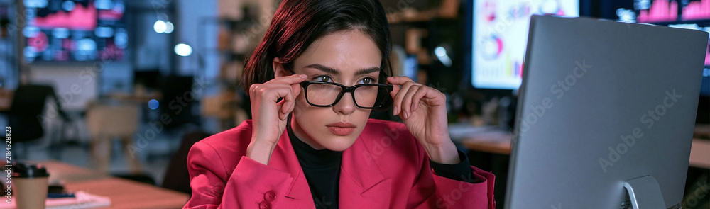 Fototapeta premium Focused Analyst: A determined woman with glasses adjusts them, intently focused on a computer screen displaying data, representing intense concentration in a professional setting.