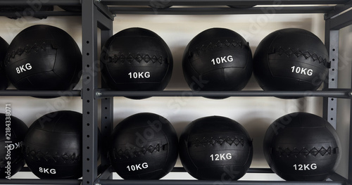 Black medicine balls of different weights arranged on a metal storage rack in a modern gym. Fitness equipment for strength training, functional workouts, CrossFit and exercise.