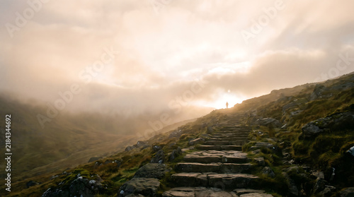 Solitary figure ascending mystical stone stairs towards a surellisme heaven in a foggy mountain landscape at sunrise.
