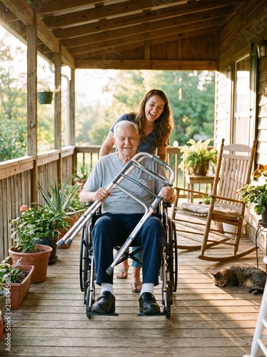 Young woman pushing senior man in wheelchair on porch with walker