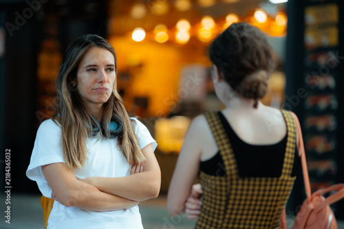 Upset Offended Woman Standing With Arms Crossed Looking At Friend. Annoyed girlfriend glares unhappily toward another girl during a tense urban conversation