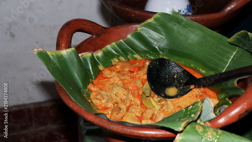 Spicy coconut milk curry inside an authentic clay pot lined with fresh banana leaf using traditional wooden ladle spoon for traditional indonesian culinary.