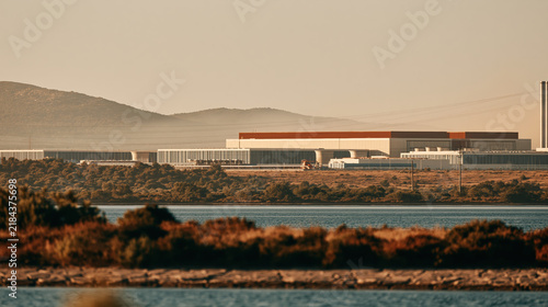 Industrial data center complex beside dry cracked ground and calm water under hazy mountain light, creating austere climate scene