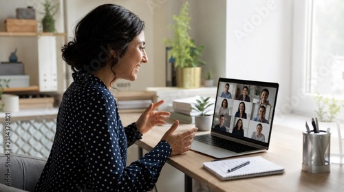 Smiling dark-haired woman video calling colleagues on laptop at bright home desk during remote meeting, gesturing while engaged in virtual conversation in sunlit office space