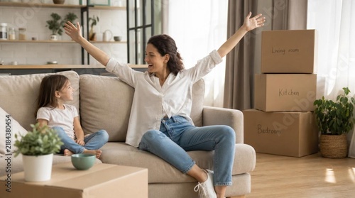 Joyful young mother raising arms in celebration while sitting on beige sofa with happy little daughter, moving day with cardboard boxes labeled living room, kitchen, bedroom in bright modern interior