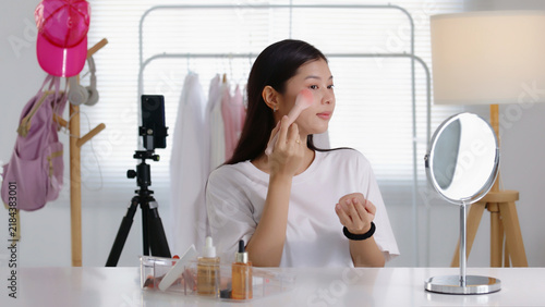 Fotografía Young Asian beauty influencer applying blush with a powder brush while recording a makeup tutorial on smartphone in her room, perfect for cosmetics, blogging, and social media content