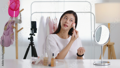 Quadro su tela Young Asian beauty influencer applying blush with a powder brush while recording a makeup tutorial on smartphone in her room, perfect for cosmetics, blogging, and social media content