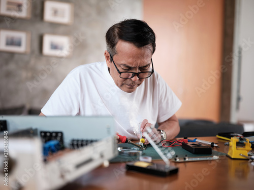 Mature Asian man repairs home electrical appliance, disassembling and soldering circuit board, focusing on precision detail, DIY fix tools, electronics component workshop hobby, expert handyman.